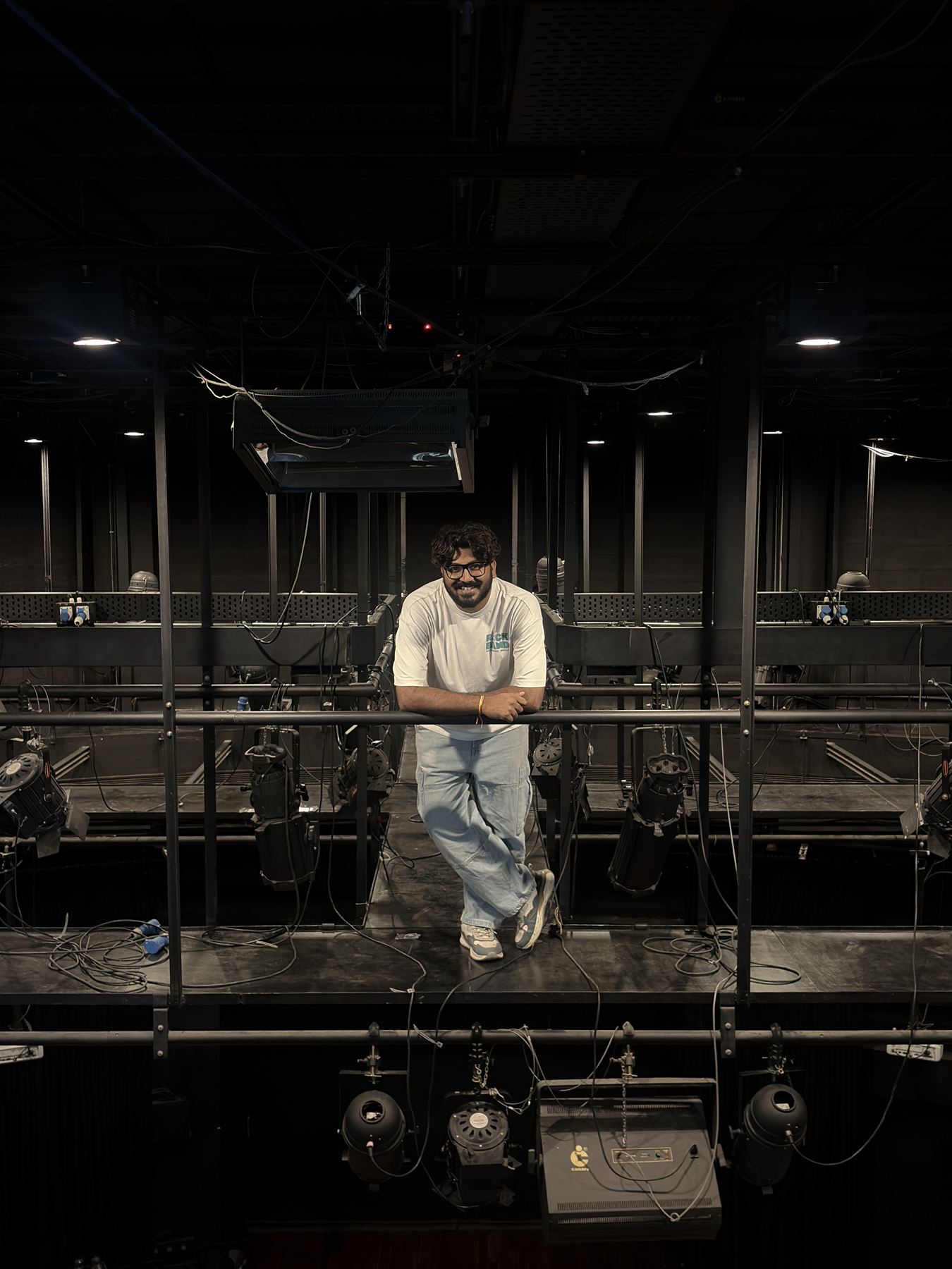Chiraag standing on the lighting grid above a theatre stage, surrounded by stage lights