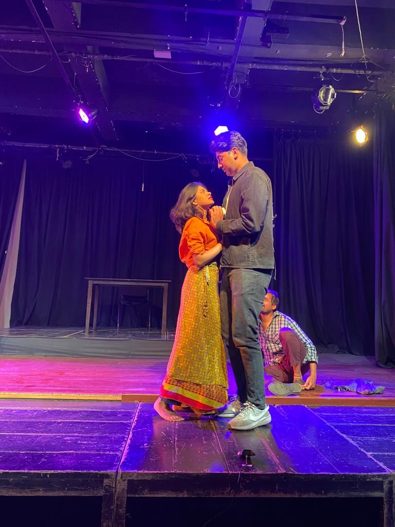 Two actors embrace on a stage washed in purple and amber light